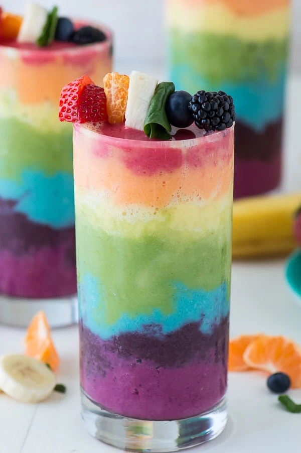 fruit kabobs in rainbow colors, topping a tall glass, filled with a rainbow colored drink, fruit and spinach smoothie 