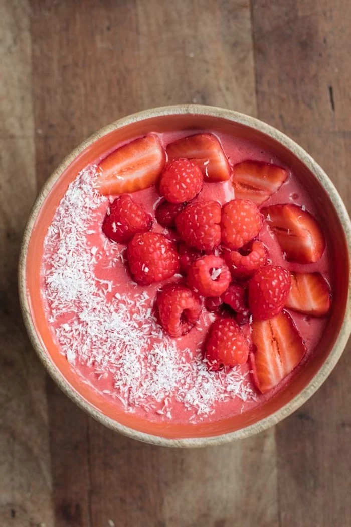 dusting of coconut shavings, topping a bowl with a pink puree, how to make a fruit smoothie, topped with whole raspberries, and strawberry slices