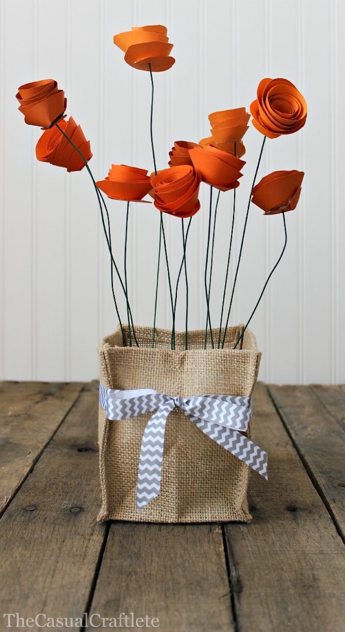 square burlap bag, decorated with grey and white ribbon, mothers day presents, containing eleven reddish paper flowers, with green wire stalks