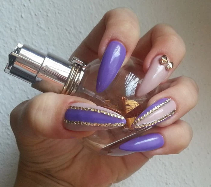 kitsch-style claw nails, in violet and light pink, decorated with lots of tiny, round golden rhinestone stickers, and a single golden bow shape, on hand-holding a perfume bottle
