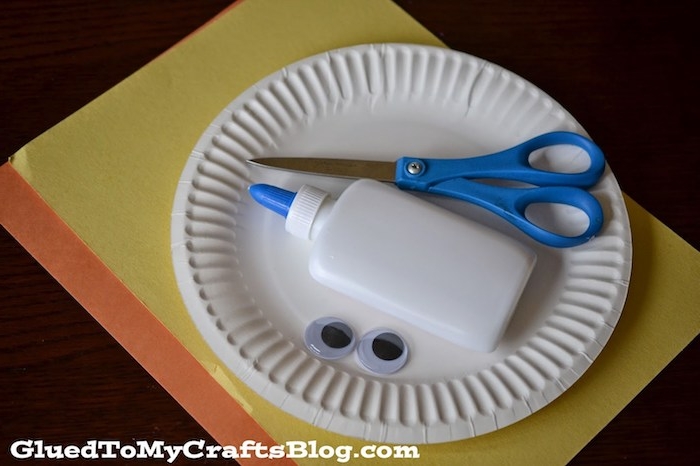 plastic glue bottle and scissors, googly eye stickers, inside white paper plate, placed on sheets of yellow, and orange card, easter crafts for preschoolers