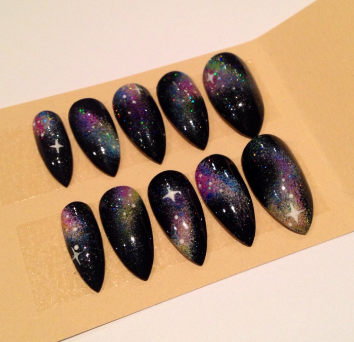 ten faux black stiletto nails, decorated with pink, blue and yellow sprays of paint, to look like the night sky, white hand-painted stars, and glitter