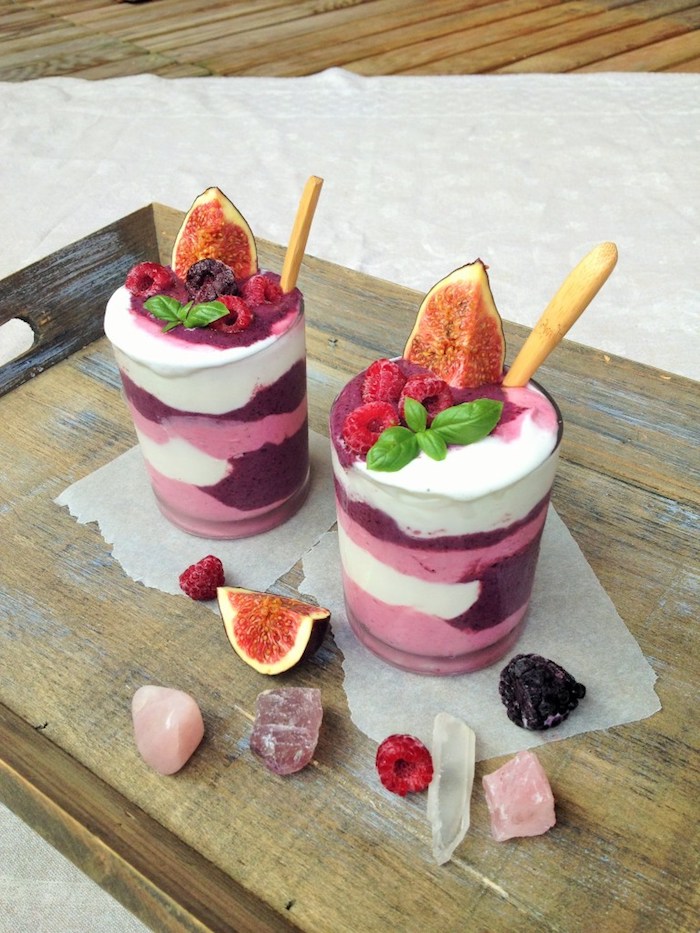 crystals and berries, and a slice of fig, near two glasses, containing pale pink, white and purple, creamy liquid, topped with fruit and mint, layered smoothie recipes