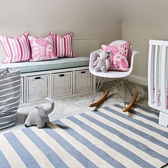 four vibrant pink and white cushions, placed on storage bench, and small white rocking chair, baby girl room décor, striped rug in cream and blue, stuffed elephant toy