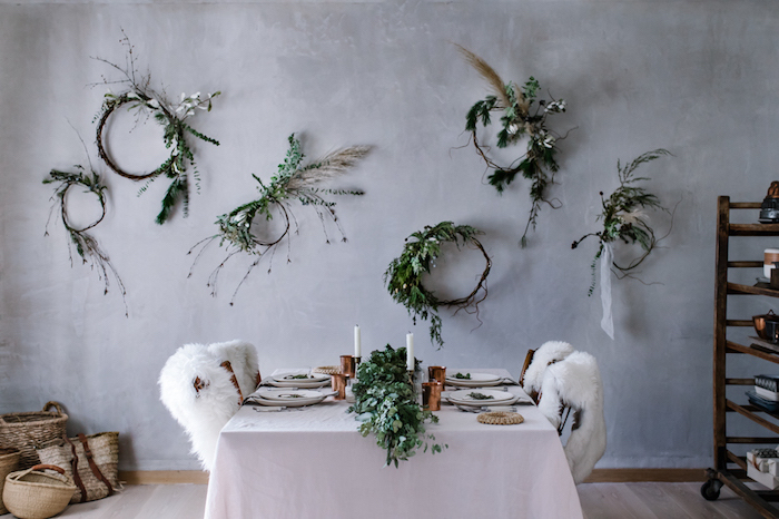 set table with white tablecloth, several wooden chairs, covered with lambskin throws, rustic country home décor, six decorative plant and flower wreaths, hung on a pale gray wall