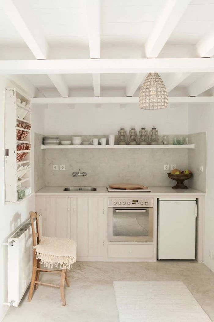 minimalist kitchen in white and ivory, with small fridge, modern stove and several cabinets, retro sink and chair, decorative plates and gas lanterns