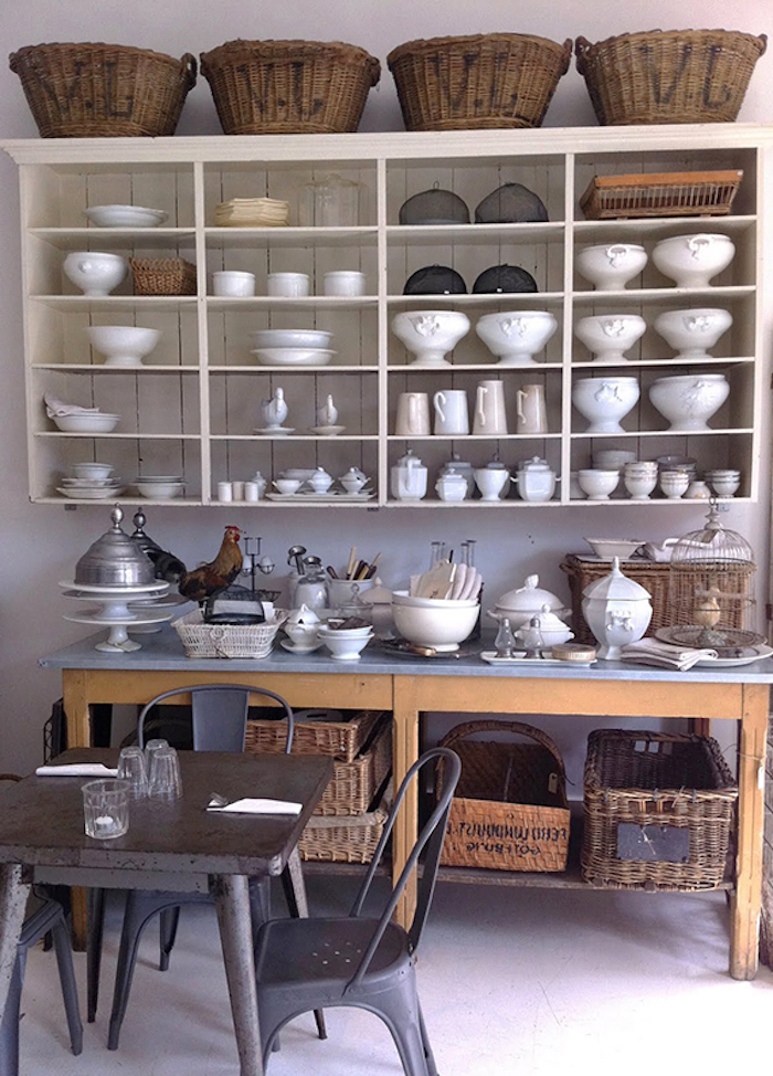 soup dishes and mugs, bowls and jugs, inside an off-white cupboard, near large wooden counter, covered with various dishes and crockery, and a small square black metal table, with matching chairs