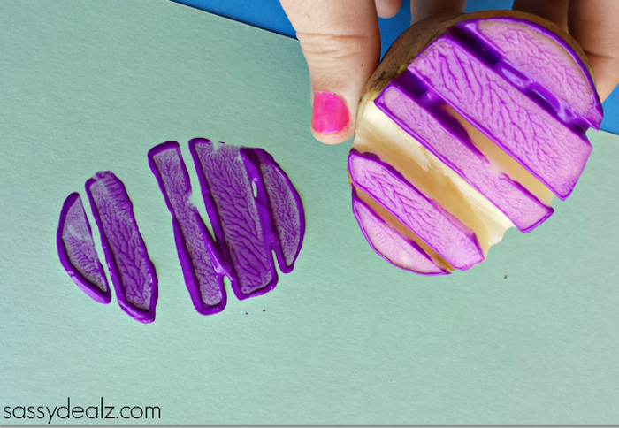 potato stamp covered in paint, held by hand with pink nail polish, easter crafts for preschoolers, sheet of pale green card, with purple easter egg-shaped print