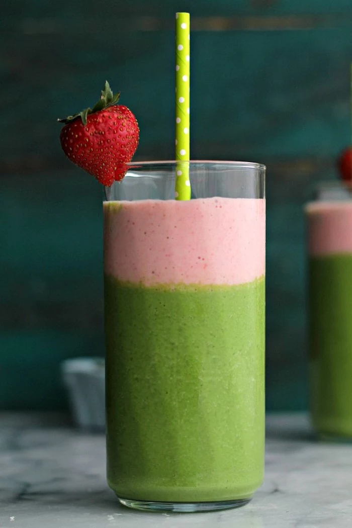 paper straw in bright green, inside a tall glass, decorated with a whole strawberry, containing a creamy layered fruit drink, pink and green smoothie recipe 