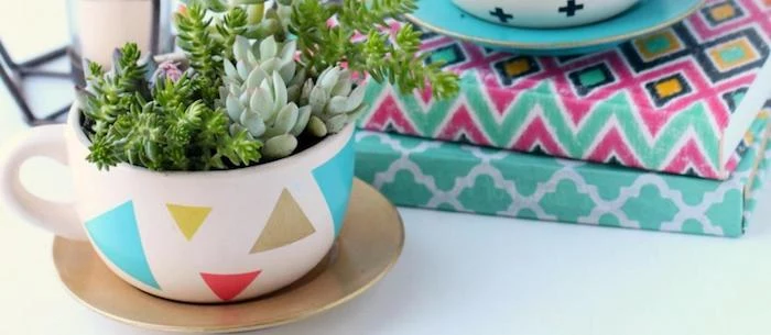 coffee cup decorated with colorful triangles, placed over a golden saucer, containing dirt and several planted succulents, good mothers day gifts, multicolored books in background