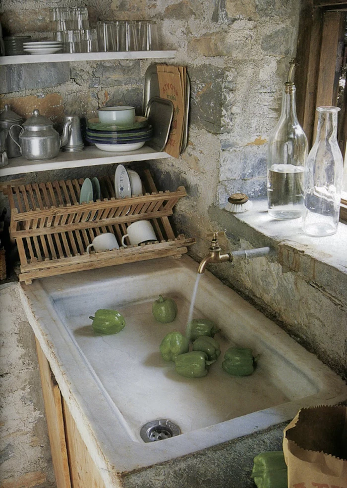 natural stone sink basin, containing seven green peppers, vintage tap with running water, country kitchen décor, stone covered walls, simple white shelves, and a wooden dish drying rack