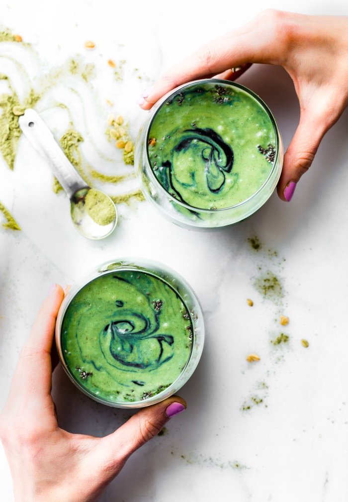 matcha powder in a green smoothie, poured into two glasses, each held by a hand, with pink nail polish, measuring spoon with matcha nearby