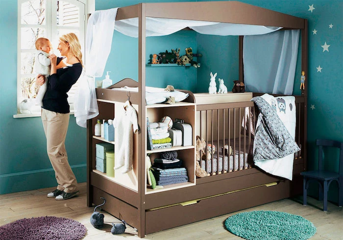 all-in-one baby crib, with changing table, storage space and shelves, made from brown wood, inside a boy nursery, with teal blue walls, blonde woman holding a baby