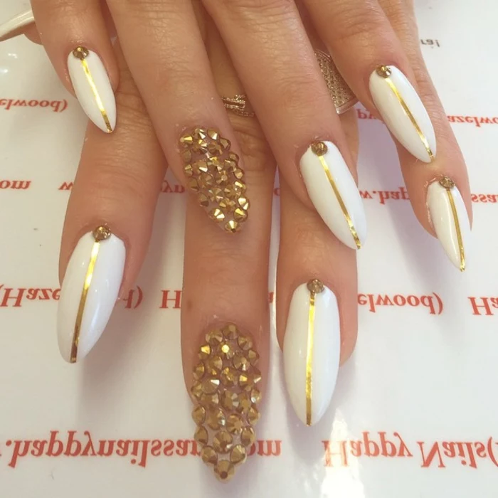 encrusted nails with gold rhinestone stickers, white nail polish, decorated with thin golden stripes, long oval and sharp manicure