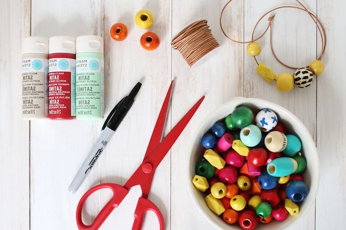 scissors in red, white bowl filled with multicolored wooden beads, mothers day gifts, three tubs of paint, thread and a pen