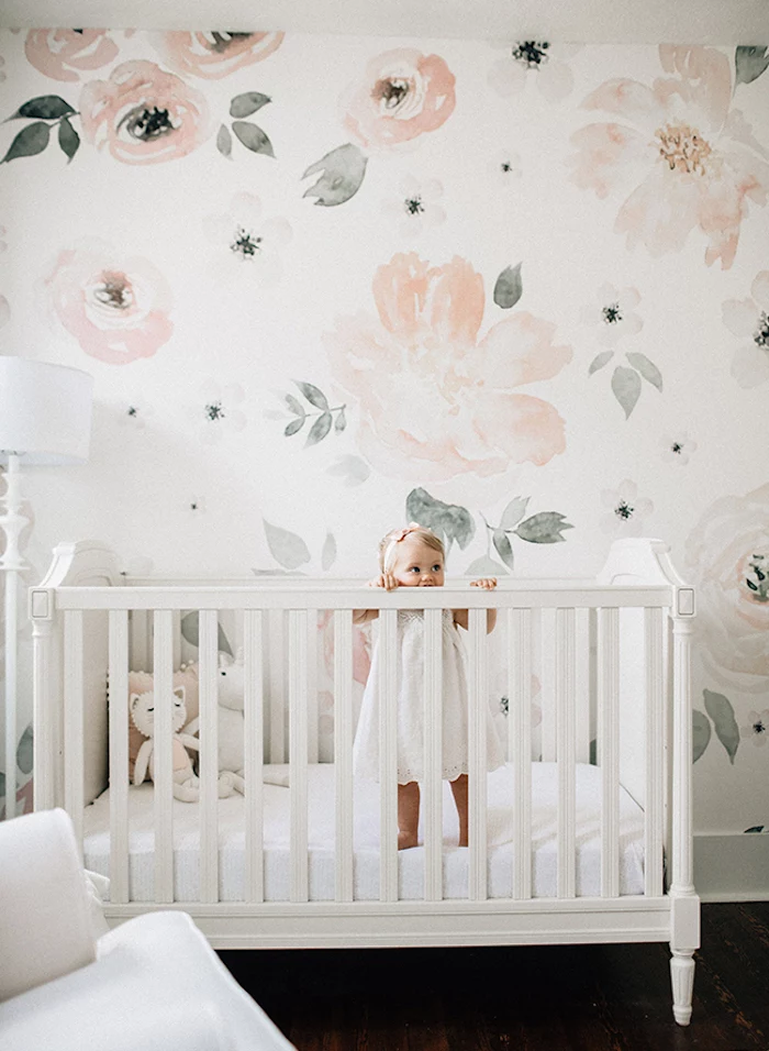 little toddler girl, standing in white, ornate wooden crib, baby girl themes, near white wall, with pale pastel, water color-like floral print