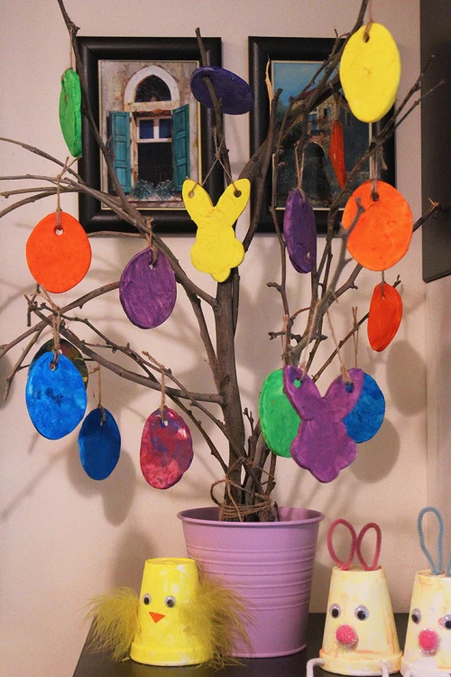 easter tree, made from dried branches, decorated with differently colored, hand-crafted and hand-painted ornaments, shaped like eggs and bunnies, easter crafts for preschoolers, more hand-made decorations nearby