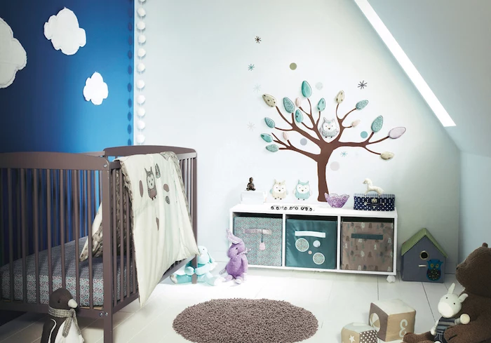 white clouds painted on a blue wall, in boy nursery, other visible wall is white, and features a tree mural, with colorful leaves and owls, brown wooden crib