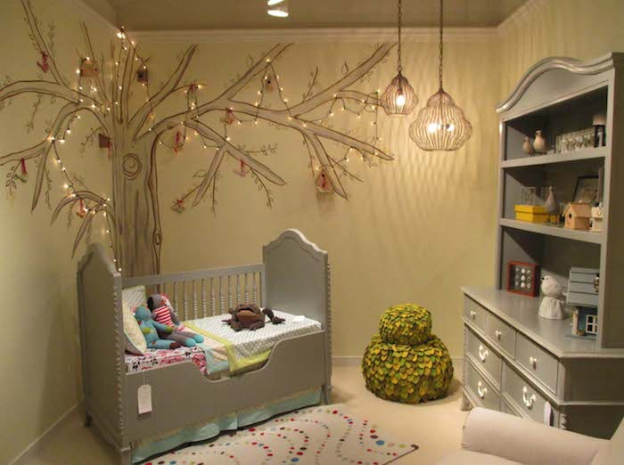 matching gray ornamental crib, and cupboard with shelves, in baby nursery, with pale yellow walls, and tree mural, decorated with tiny fairy lights