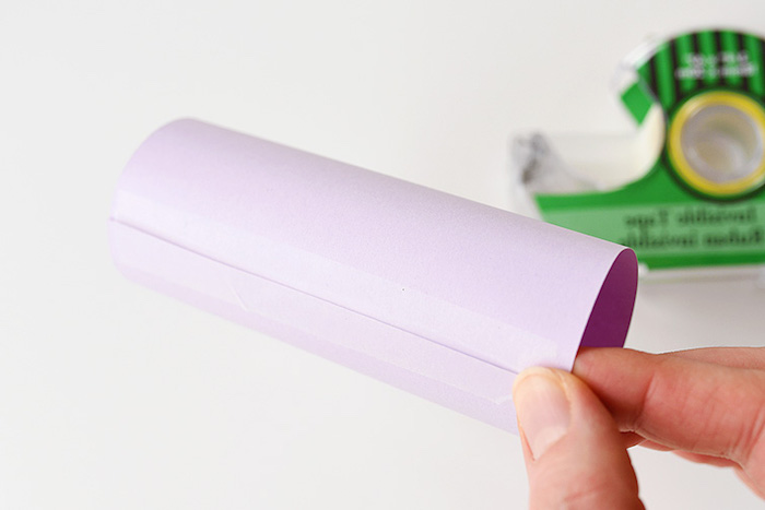 tube made out of pale, rolled purple card, stuck with clear sticky tape, and held by a hand, easter diy, sticky tape dispenser in background
