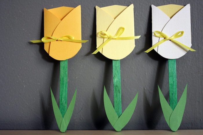 kid's crafts projects, mother's day gifts for grandma, three paper tulips, in yellow and white, with small ribbons, green paper leaves, and hand-colored stalks