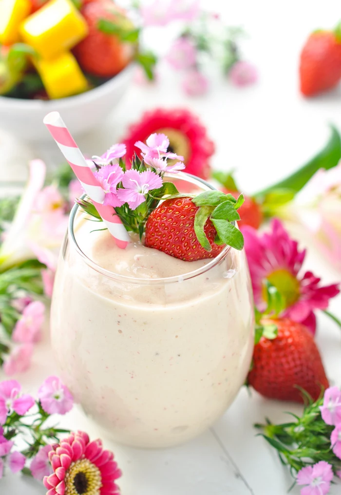 strawberry and small pink flowers, topping a tumbler glass, filled with pale pinkish-beige drink, protein shake recipes, more berries and flowers nearby