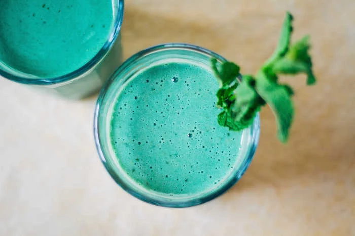 turquoise frothy liquid, blue-green smoothie recipe, poured in two identical, tall clear glasses, one topped with a green plant