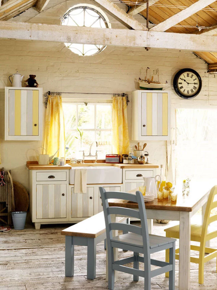 chairs in pastel blue and yellow, near small square wooden table and bench, inside a room with white brick walls, ceiling beams and wooden floor, pale grey and white, striped country kitchen cabinets