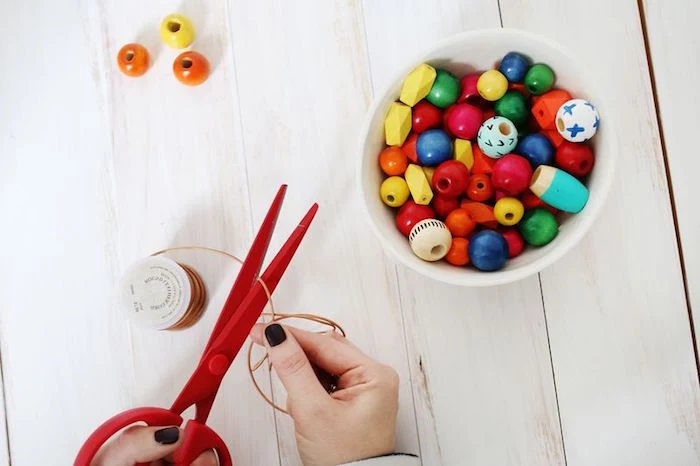 mother's day gift ideas, cutting leather cord, with pair of red scissors, white bowl filled with multicolored, wooden bead nearby