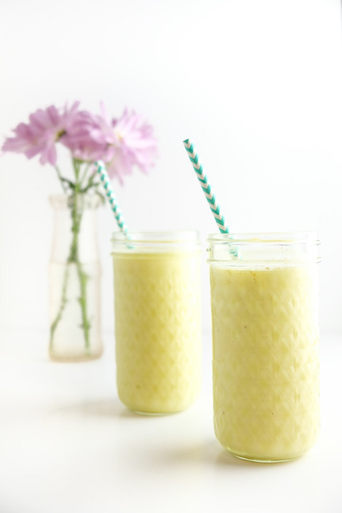 purple flowers in a clear vase, near two tall glasses, containing a pale yellow blended drink, healthy smoothie recipes, two striped straws