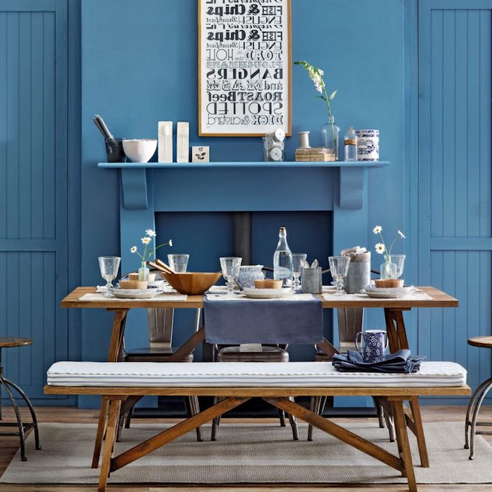 vivid blue walls, and a fireplace, inside a dining room, with light brown wooden table, matching bench and chairs, country kitchen décor, bowls and plates, mugs and glasses