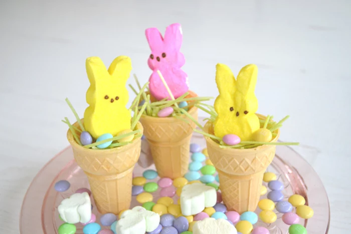 peeps shaped like bunnies, in yellow and pink, placed in light wafer cones, with green easter glass and candy, on plate with marshmallows, and other sweets