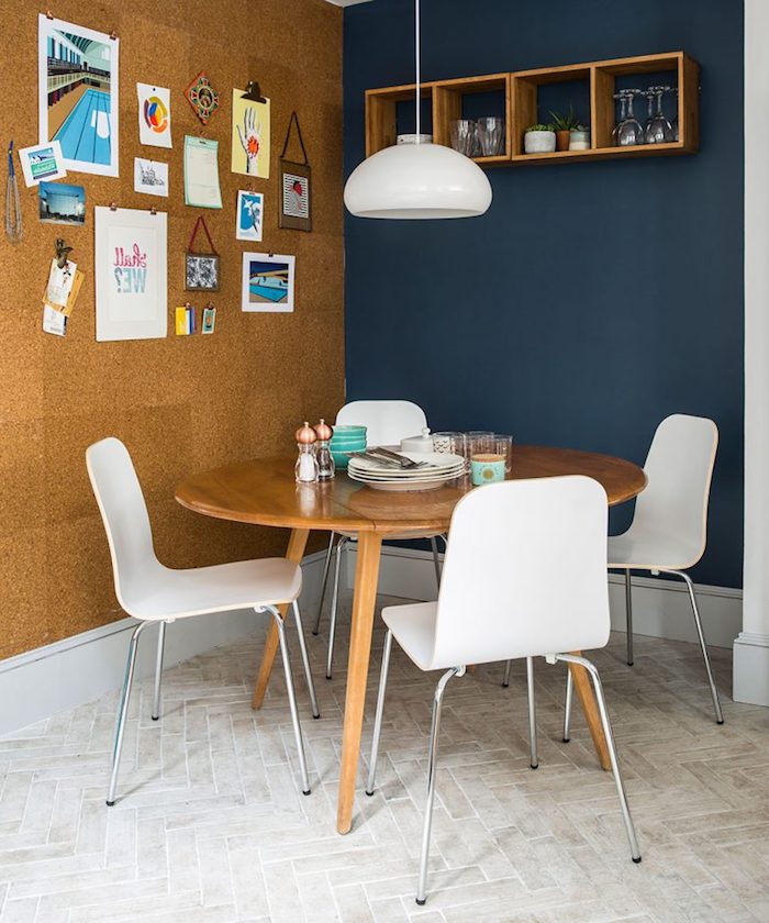contrasting walls in navy and beige, one with four square wooden shelves, the other decorated with various photos, images and drawings, country kitchen décor, round wooden table, with four modern white chairs
