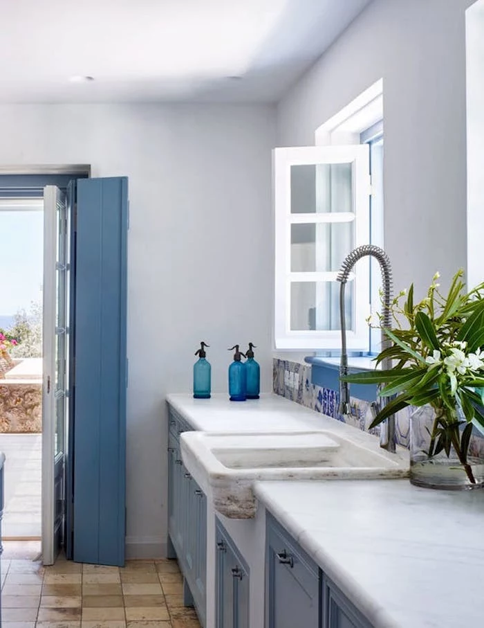 pale blue rustic kitchen cabinets, inside white room, with stone sink, and modern metal water tap, opened window and door, floor with beige stone tiles