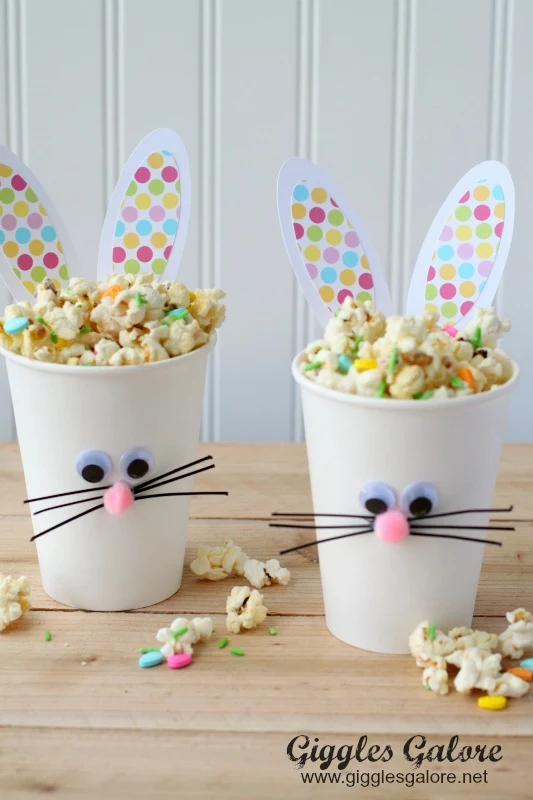 cups made from white paper, decorated with rabbit ears, googly eye stickers, pom pom noses, and black fuzzy wire whiskers, containing multicolored popcorn, great easter idea