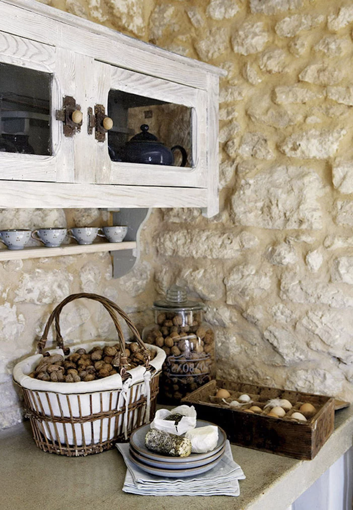 walnuts inside a basket and a large jar, near wooden box filled with eggs, and several gray plates, sandy-yellow stone wall, pale gray rustic cupboard