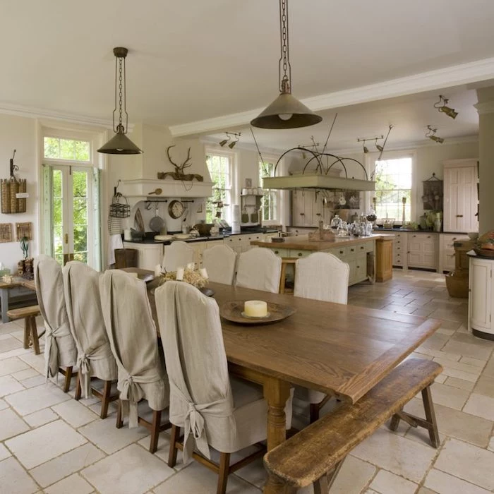 spacious kitchen with large dining table, worn wooden bench, and four chairs covered in beige cloth, rustic country home décor, white cupboards and appliances, various decorative objects