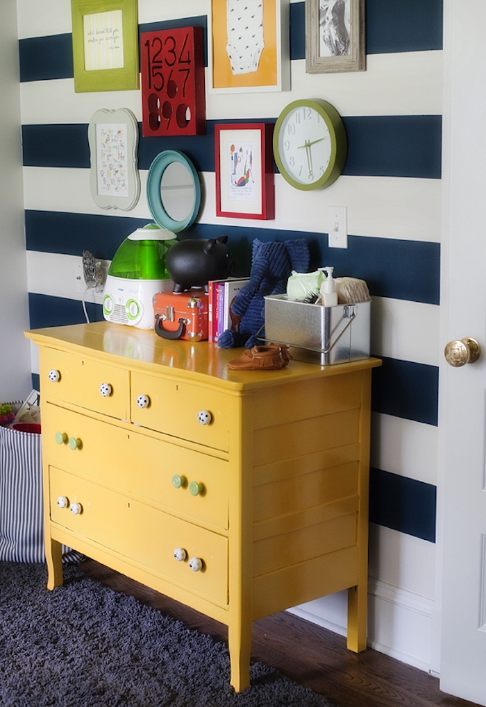 chest of drawers in bright yellow, covered with baby items and toys, nursery ideas, next to white wall, with navy stripes, decorated with images in colorful frames, mirror and a wall clock