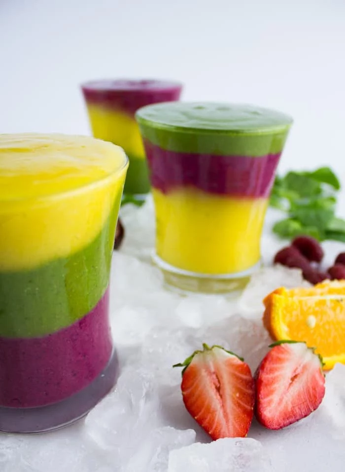 layered blended fruit drinks, purple yellow and green smoothie, in three glasses, near pieces of strawberry, orange and mint