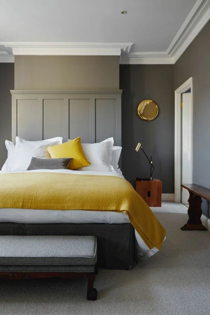 bench in dark brown, creamy grey bedroom with paneling, light gray carpet, bed with white and grey, and yellow covers and pillows