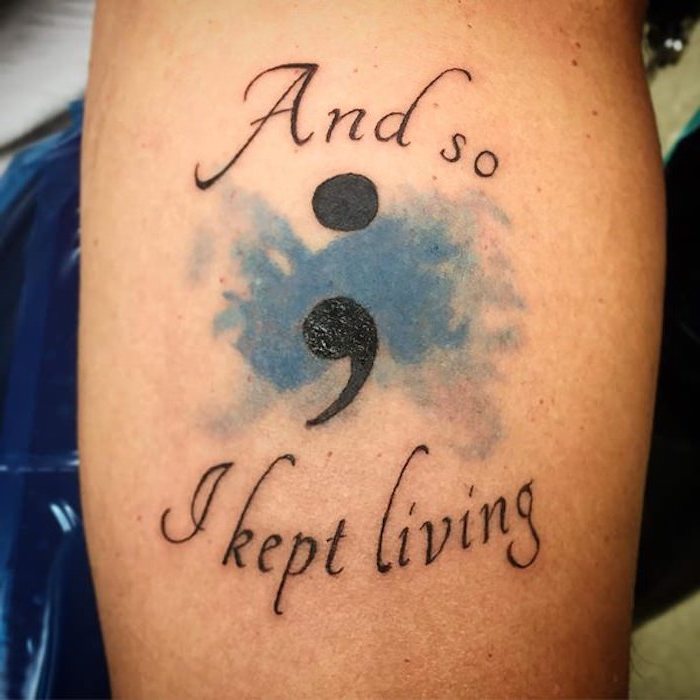 large semicolon in black, tattooed with the message and so i kept living, also in black with a splash of watercolor-like blue, semicolon tattoo meaning, on person's arm