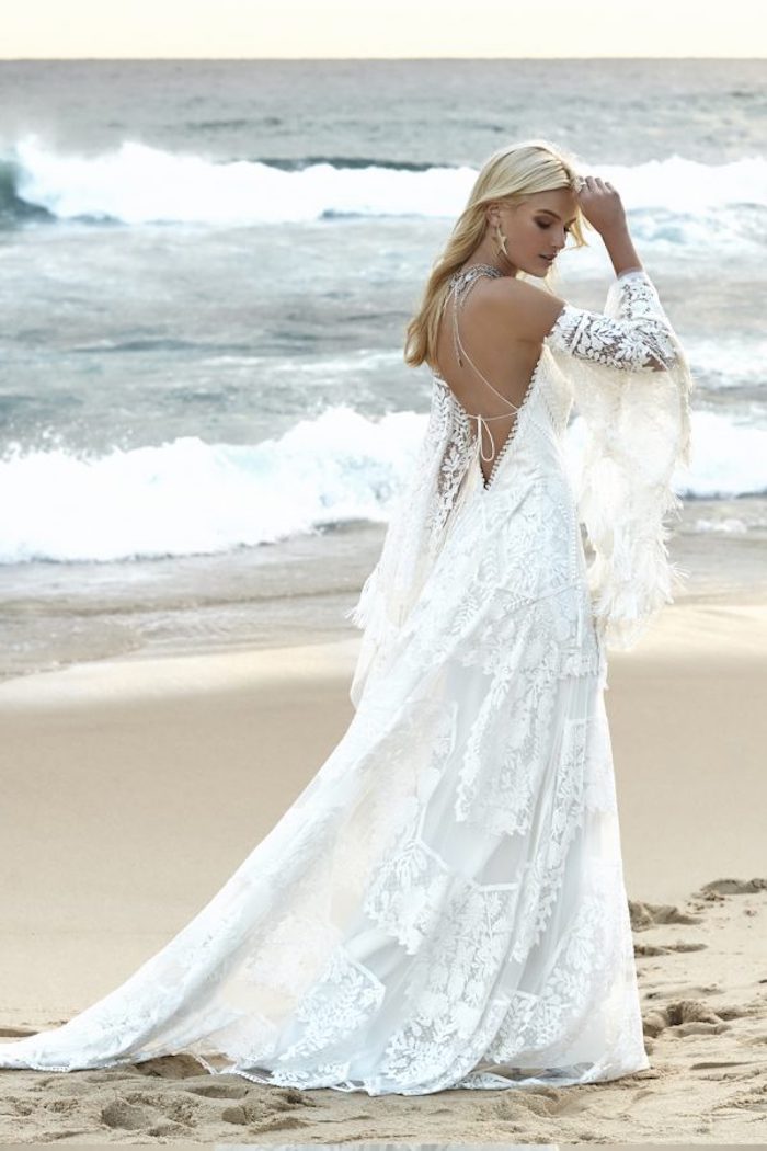boho lace beach wedding dress, in white with wide tasseled sleeves, and a cutout back, worn by blonde woman, walking on a sandy shore