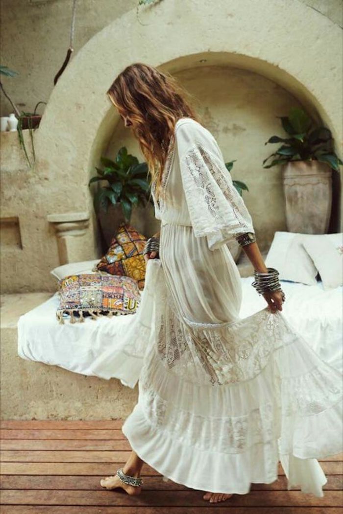 stonewall with arch and an inbuilt bench, covered with a white blanket and multi-colored cushions, and decorated with green potted plants, woman in boho maxi dress, with lace and embroidery, walking barefoot while holding her skirt