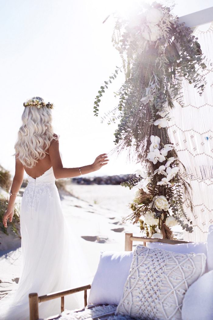 flower crown on a platinum blonde, curled long hair, worn by tanned slim woman, in a backless white gown, wedding dresses for beach wedding, settee with cushions and flowers nearby