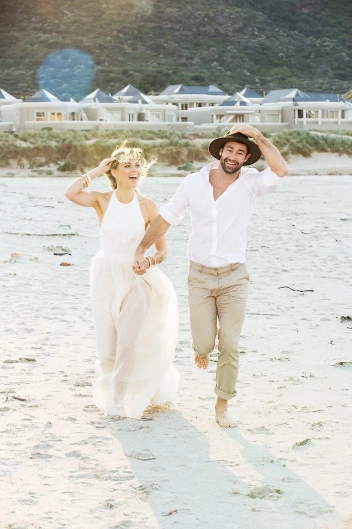 semi-sheer white sleeveless gown, casual beach wedding dresses, worn by laughing blonde bride, with flower crown, running on a beach, hand in hand with a groom, in white shirt, beige pants and a hat