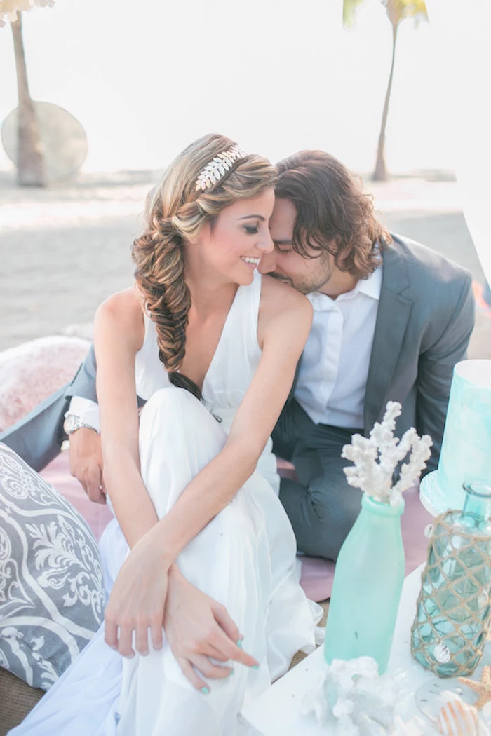 intimate image of a groom, wearing grey suit and white shirt, sitting next to a bride, in a long white gown, and kissing her shoulder, wedding dresses for beach wedding, marine-inspired decorations and cushions