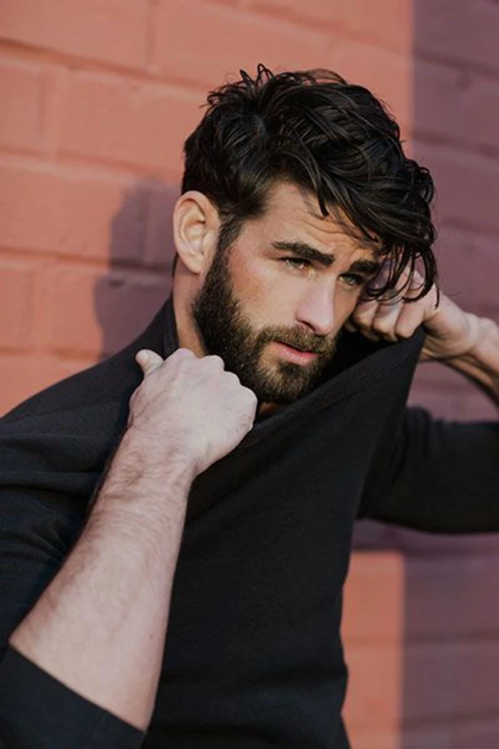 man pulling his black jumper with both hands, with black shiny hair, wavy and layered, haircuts for curly hair, beard and mustache, thick bushy eyebrows
