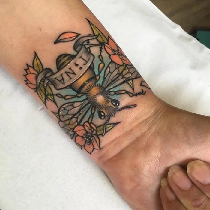 wrist tattoo featuring a large bee, several orange flowers, with green leaves and thick black outlines, and a banner with the name tina written in black, the i substituted for a semicolon tattoo 