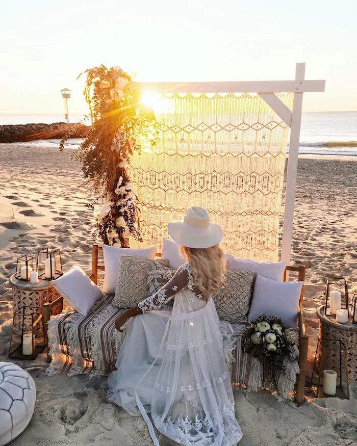 beach weddings in florida, boho decor on a sandy shore, bride in long dress and hat, and sheer white embroidered maxi cardigan, sitting on a bench covered in cushions, and watching the sunset