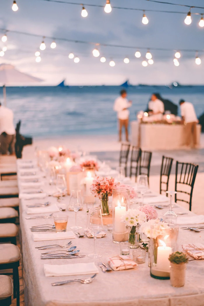 long set table, with cutlery and plates, glasses and candles, small flower bouquets in white and pink, on a beach near the sea, florida destination weddings, two rows of string lights